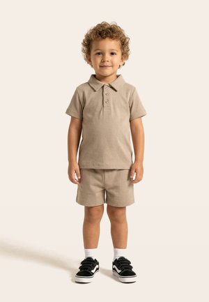 Bambino piccolo con capelli ricci che indossa una polo beige e pantaloncini, calzini bianchi e scarpe da ginnastica nere, in piedi e rivolto in avanti.