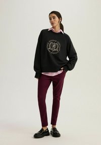 Czarna bluza z złotym emblematem, nałożona na jasnoróżową koszulę w paski. Bordowe spodnie slim-fit i czarne skórzane buty dopełniają całości.