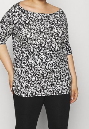 Kobieta ubrana w czarno-biały top w kwiatowy wzór z rękawami do łokcia i czarne spodnie, stojąca na jednolitym tle.