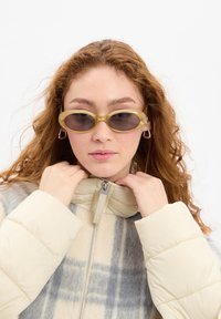 Sunglasses with a thin gold frame and dark lenses, worn with a light beige, puffy, zip-up jacket featuring a gray and blue plaid pattern.