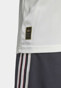 Hvit mesh idretts skjorte med et teksturert mønster, med et svart og gyllent "Authentic Licensed Product"-merke, sammen med mørke shorts med tre striper.