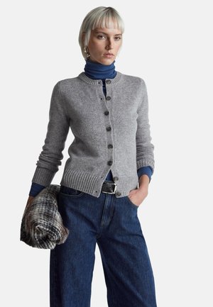 Graue Wollstrickjacke mit Rundhalsausschnitt, Knopfleiste vorn, geripptem Saum, kombiniert mit einem blauen Rollkragenpullover und hoch taillierter Jeans.