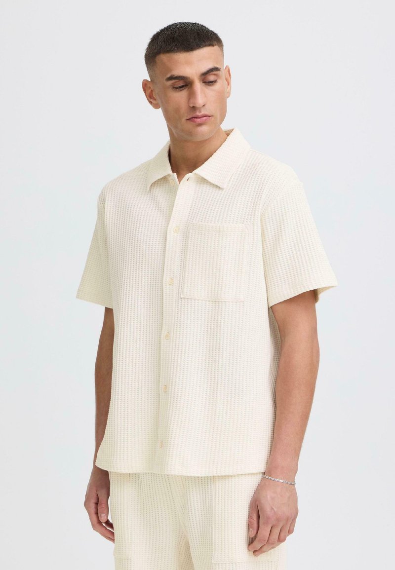 Jeune homme portant une chemise à manches courtes texturée couleur crème avec poche frontale, regardant vers le bas, debout devant un fond clair uni.