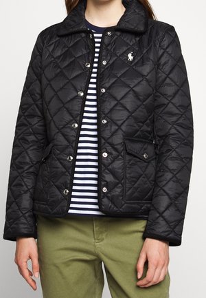 Personne portant une veste matelassée noire avec fermeture par boutons, poches avant et un petit logo blanc sur la poitrine, par-dessus une chemise rayée et un pantalon vert.