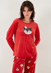 Rotes Langarmoberteil mit einem Grafik eines schwarzen Katers und Blumendetails; kombiniert mit roten Hosen mit Tiermustern und verspieltem Text.