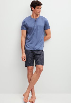 Man met een blauw henley-shirt met korte mouwen en donkere korte broek met patroon, staand op blote voeten tegen een witte muur, kijkt opzij.