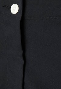 Czarna tkanina o gładkiej strukturze, z pojedynczym białym guzikiem i subtelnymi detalami przeszyć. Skup się na guzikach i jakości materiału.