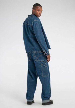 Mann, der rückwärts steht, Kopf gedreht, trägt eine locker sitzende blaue Jeansjacke und Jeanshose mit schwarzen Schuhen vor einfachem Hintergrund.