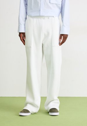 Man wearing loose white pants with front pockets and green slip-on shoes, standing on light green floor against a plain white wall.