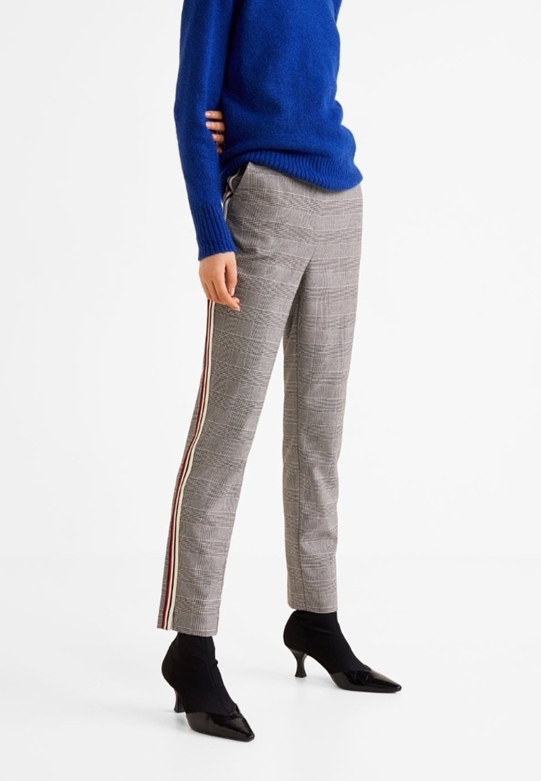 Personne portant un pantalon à carreaux gris avec des bandes latérales rouges et blanches, un pull bleu et des chaussures à talons noires, debout devant un fond blanc.