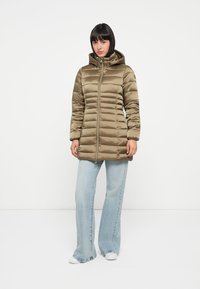 Olivengroene pufferjack met een capuchon, voorzien van gequiltte secties, een ritssluiting aan de voorkant en een iets langere snit. Gecombineerd met lichtblauwe jeans.