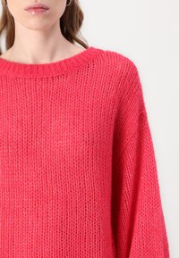 Pull en maille rose avec un col large à côtes et des manches à coupe ample. La texture est douce et épaisse, avec un design légèrement transparent.