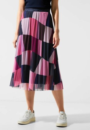 Woman wearing pleated midi skirt with abstract pink, purple, and black pattern paired with white sneakers and a black top.