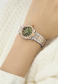 Rožu zelta un sudraba metāla pulkstenis ar astoņstūru korpusu, zaļu ciparnīcu, dimanta līdzīga noformējuma akcentiem un saistītu jostu uz bēša plaukstas.