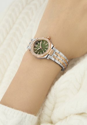 Złoty róż i srebrny metalowy zegarek z ośmiokątną kopertą, zieloną tarczą, diamentowymi akcentami i ogniwowym paskiem na beżowym nadgarstku.