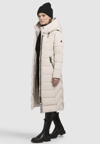 Lang dunjakke i lyse beige med høj krave, chevron kviltet mønster, frontlynlås og sorte akcentdetaljer. Båret med sorte støvler.