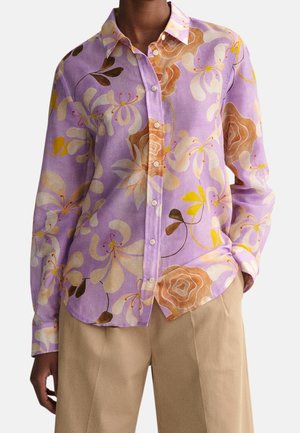 Chemise violette boutonnée avec de grands motifs floraux beige, marron et jaune, portée avec un pantalon beige, main dans la poche droite.