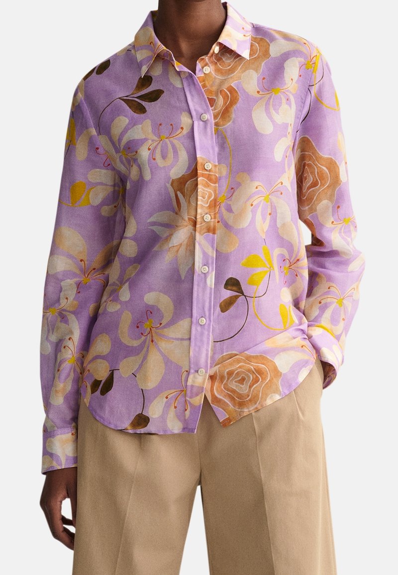Chemise violette boutonnée avec de grands motifs floraux beige, marron et jaune, portée avec un pantalon beige, main dans la poche droite.