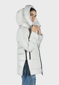 Chaqueta de plumón de color gris claro con cuello alto y capucha grande, que cuenta con bolsillos laterales con cremallera, un diseño aerodinámico y secciones cosidas para aislamiento.