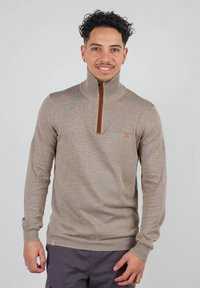 Pull en tricot beige avec un col zippé, featuring une fermeture éclair marron et un petit accent de logo. Coupe slim avec des poignets et un ourlet côtelés.