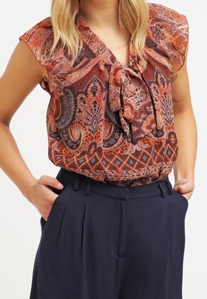 Femme portant un chemisier à motif cachemire rouille et noir avec un nœud à l'avant, assorti à un pantalon plissé bleu marine taille haute, les mains dans les poches.