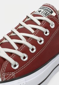 Rdeča platnena superga z belimi vezalkami, srebrnimi sredicami in gumijastim podplatom. Ima črno-belo logotipno nalepko na zunanji strani.