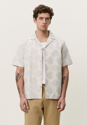 Mann mit lockigem Haar und Tattoos trägt ein beige gemustertes kurzärmeliges Hemd über einem weißen T-Shirt und beiger Hose, steht vor einem einfarbigen Hintergrund.