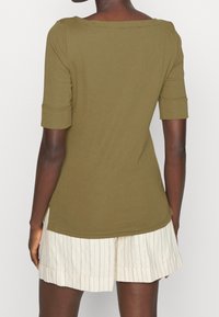 Oliivivihreä, vartalonmyötäinen paita, jossa on venepääntie ja kierretyt lyhyet hihat; valmistettu pehmeästä kankaasta, jossa on sivuaukot ja kaareva helma.