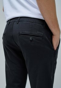 Persona che indossa pantaloni Salsa Jeans neri con tasche posteriori con bottoni e mano nella tasca destra, abbinati a una camicia bianca.