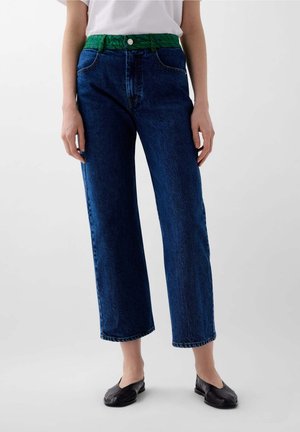 High-waisted blue denim jeans with a green patterned waistband, straight-leg design, and subtle stitching details. Paired with black flats.