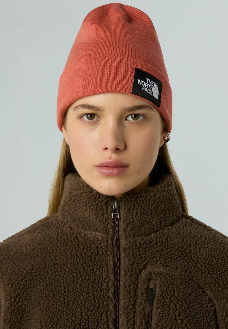 Gorro de punto naranja con un parche de logo negro, combinado con una chaqueta de forro polar marrón que tiene un cuello alto y cierre de cremallera.