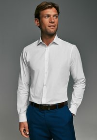 Cămașă albă cu mâneci lungi, căptușită cu guler deschis, asortată cu pantaloni bleumarin. Țesătura pare netedă și bine ajustată.