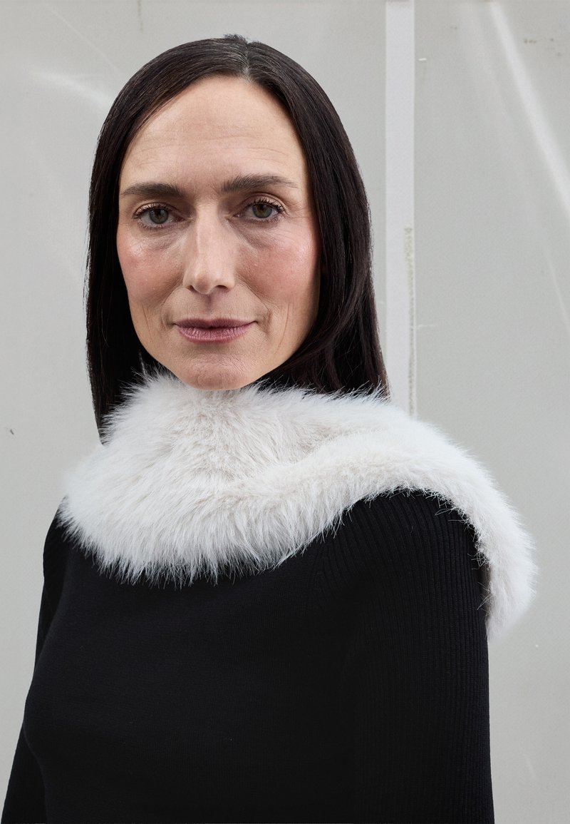 Fehér műszőrme gallér öleli körbe a nyakat, amely egy fekete bordázott pulóverhez van rögzítve. A szőrme bolyhos tapintású, puha fényű.