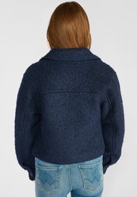 Veste en mélange de laine bleu marine avec une surface texturée, un large col et des épaules tombantes. Associée à un jean bleu clair avec un design classique de poche arrière.