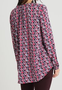 Blouse à manches longues avec un motif géométrique rouge, rose, bleu et blanc, dotée d'un ourlet arrondi et d'un détail de pincement à l'arrière.