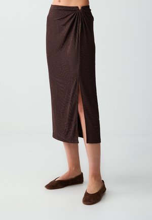 Femme portant une jupe texturée marron foncé mi-mollet avec une fente à l'avant et un détail noué, associée à des chaussures plates marron assorties.