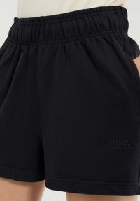 Bermudas negras con cintura elástica y bolsillos laterales, con un sutil logo bordado de ellesse en la pierna izquierda, llevadas con una parte superior de color claro.