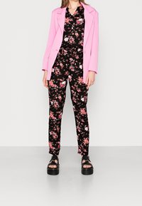 Pink blazer over a black floral jumpsuit with pink and white flowers; model wears black platform sandals; smooth fabric and tailored fit.