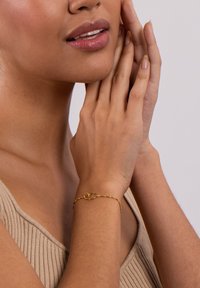 Goldarmband mit einem verschlungenen Design, bestehend aus einem ovalen Glied und einer strukturierten Kette, getragen am Handgelenk eines Modells. Der Hintergrund ist neutral.