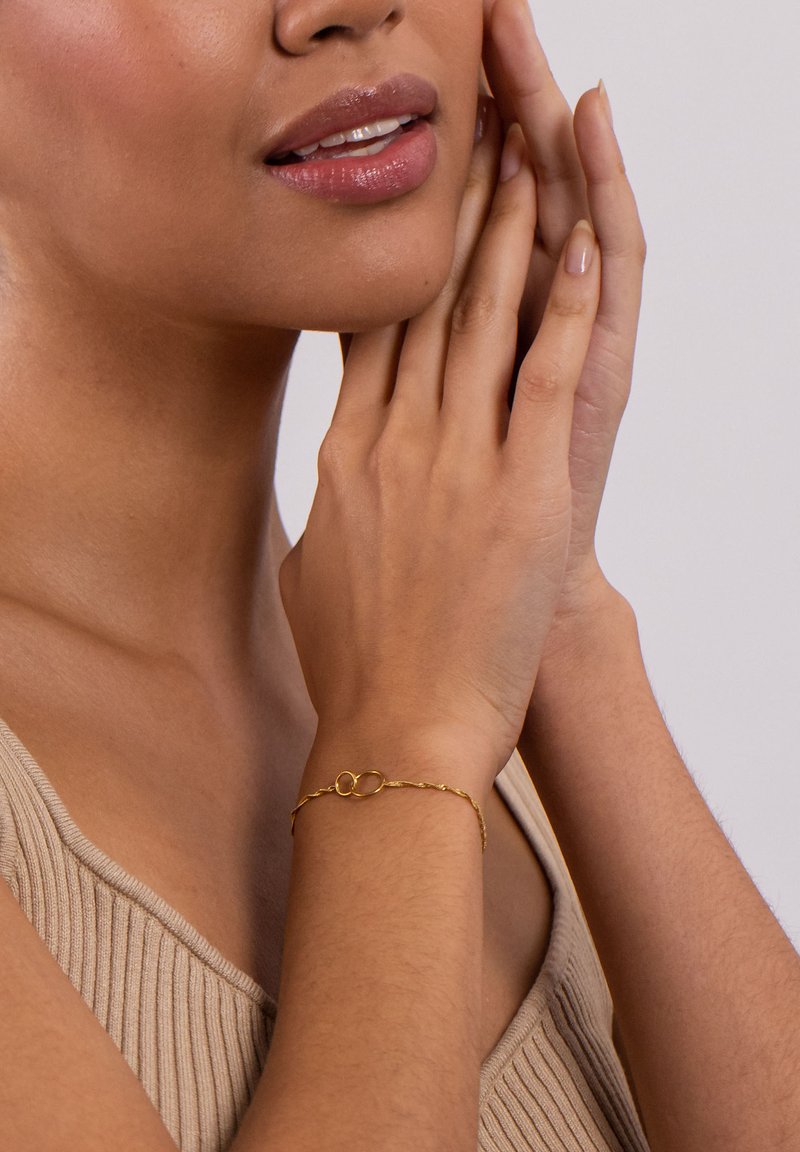 Goldarmband mit einem verschlungenen Design, bestehend aus einem ovalen Glied und einer strukturierten Kette, getragen am Handgelenk eines Modells. Der Hintergrund ist neutral.