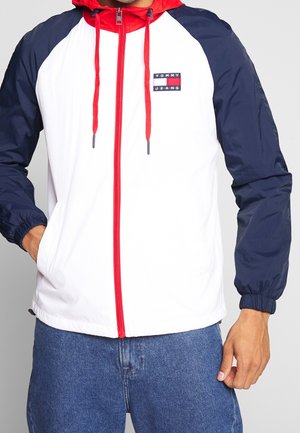 Homme portant une veste blanche avec des manches bleu marine et une capuche rouge, une fermeture éclair et des cordons, associée à un jean bleu.