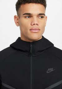 Melna zip-up kapuce ar augstu apkakli, ar tumši pelēkiem akcentiem un mazu Nike zīmodziņu uz krūtīm. Mīksta auduma tekstūra.