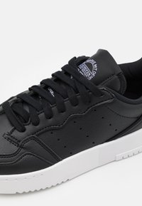 Svarta låga sneakers med texturerat läder, svarta snören, små perforeringar på sidan och en vit gummisula med greppmönster.