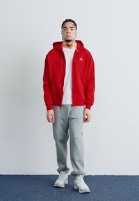 Veste à capuche zippée rouge avec cordons de serrage et logo, associée à un pantalon de survêtement gris clair et des baskets blanches, sur fond uni.