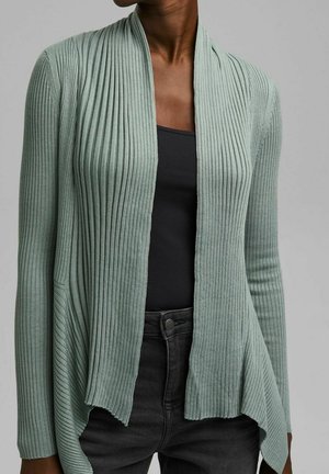 Femme portant un cardigan côtelé vert clair ouvert sur un haut noir et un jean foncé sur fond gris.