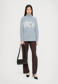 Pulover bleu cu guler înalt, având inscriptions "ROCK" în litere albe desfăcute, asociat cu pantaloni maro evazați și o geantă neagră. Texturi fine și design simplu.