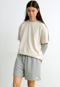 Tricou bej cu guler rotund, purtat peste o cămașă cu mânecă lungă albă și gri cu dungi, asortat cu pantaloni scurți de atletism gri, cu un logo.