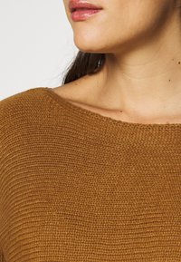 Brun strikkegenser med bred, båtformet hals, med en teksturert, ribbestrikket design. Stoffet ser mykt og koselig ut, egnet for kjølig vær.