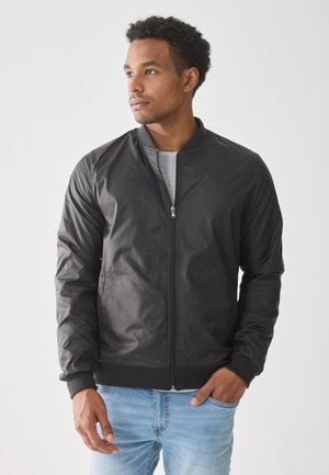 Chaqueta bomber negra con cremallera, cuello y puños acanalados. Tejido texturizado, diseño aerodinámico, usada sobre una camiseta gris con jeans de color azul claro.