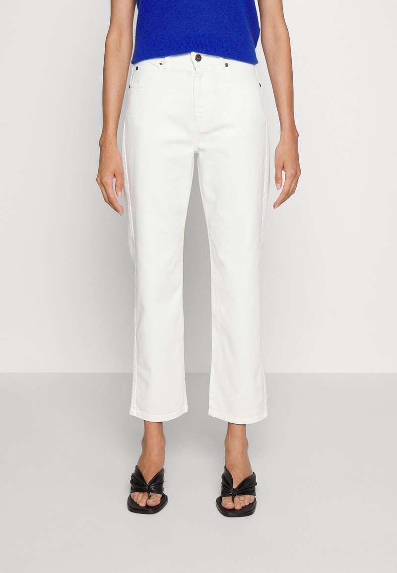 Personne portant un jean blanc taille haute coupe droite, un haut bleu et des sandales à talons ouvertes noires, debout devant un fond uni.